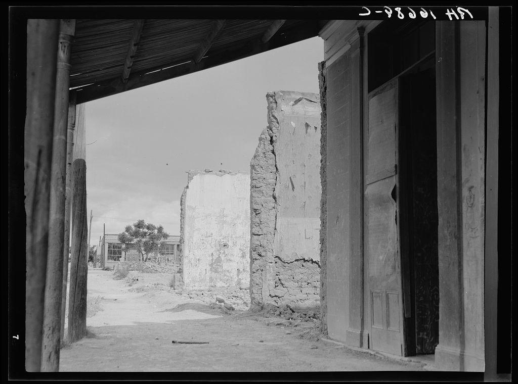 A black and white photograph depicts an abandoned urban setting. The scene is viewed from a covered walkway, with wooden beams supporting the roof on both sides of what appears to be a dirt pathway or alley. On either side of this passageway stand dilapidated structures; one shows significant damage where its wall has collapsed inward in places and another features a large gap between two walls leading into an empty interior space.

In front of these buildings, beyond the walkway's opening, lies more desolation with cracked ground and sparse vegetation. A couple of indistinct single-story buildings are visible further down this path, suggesting some level of habitation or use in what was once a community area. The absence of people adds to the sense that this place has been long abandoned.

The photograph is labeled "D-78021 H&N" at its top right corner and appears historical given the black-and-white format and style, suggesting it might have been taken during an earlier part of the 20th century. The image evokes a sense of abandonment or decay in what was perhaps once a more vibrant locality.

Additional context for this photo is provided by the URL mentioned within the description: "Scene in Tombstone, Arizona" indicates that this photograph captures a specific location known as Tombstone, which has historical significance related to mining and its boom-and-bust cycles [...]