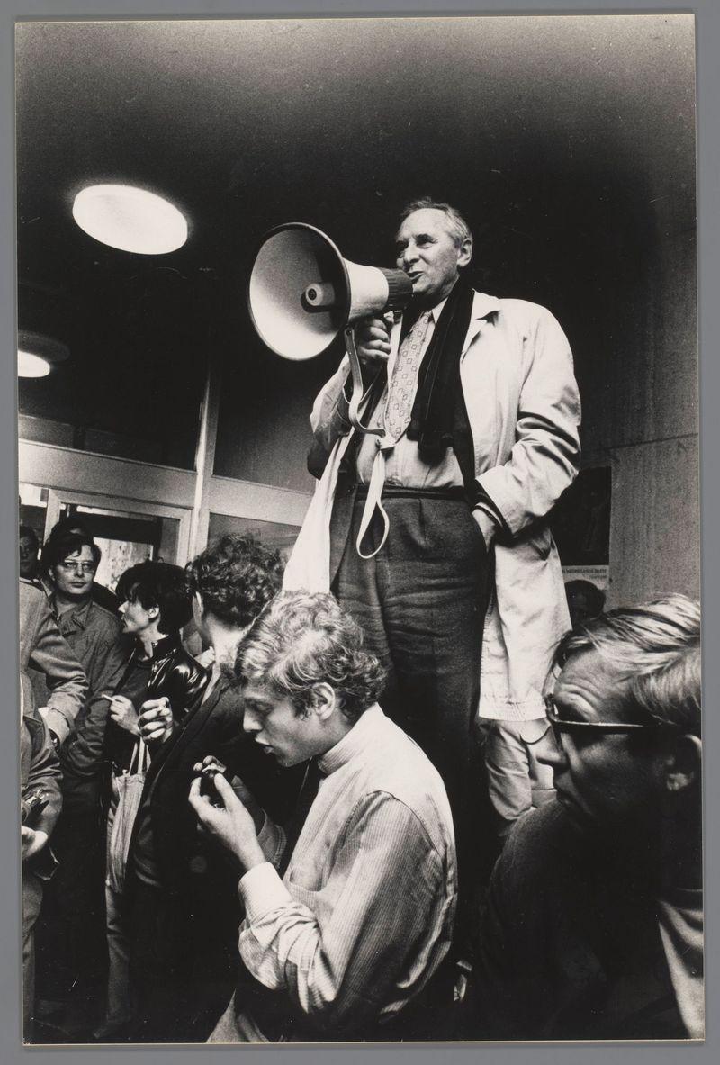 This black and white photograph captures a moment of public address. A man stands prominently in the center, elevated on what appears to be a platform or step ladder. He holds a megaphone close to his mouth with both hands, suggesting he is addressing an audience out of frame. His attire includes a light-colored jacket over a dark shirt and pants, paired with glasses resting atop his head.

Surrounding him are several individuals who seem engaged in various activities; some appear to be looking at their own devices or interacting among themselves. The lighting focuses on the central figure, creating contrast that highlights both him and those directly surrounding him against a darker background. The setting seems informal but is likely an indoor space given the ceiling lights visible above.

The image has a documentary feel, potentially documenting a specific event captured by Ed van der Elsken in Amsterdam during 1969 as indicated by the provided context link.