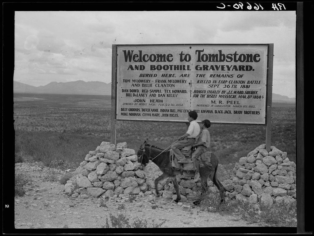 A black and white photograph of a sign with two people on horseback passing by. The text on the sign welcomes visitors to Tombstone, Arizona's Boothill Cemetery Graveyard. It lists various individuals who are buried at this site or whose remains were located here including notable figures from history such as Frank McIveye (killed in Earp-Clanton battle), Bill Davidson and Red Samuels (lynched by law enforcement for the Bisbee Massacre). The photograph is labeled with "O-P-O-W-S-H-A" at the top right corner, indicating it may be part of a series or collection. Additional information about this image can also be found in another source mentioned as Sign entering Tombstone, Arizona on the website images.loener.nl.