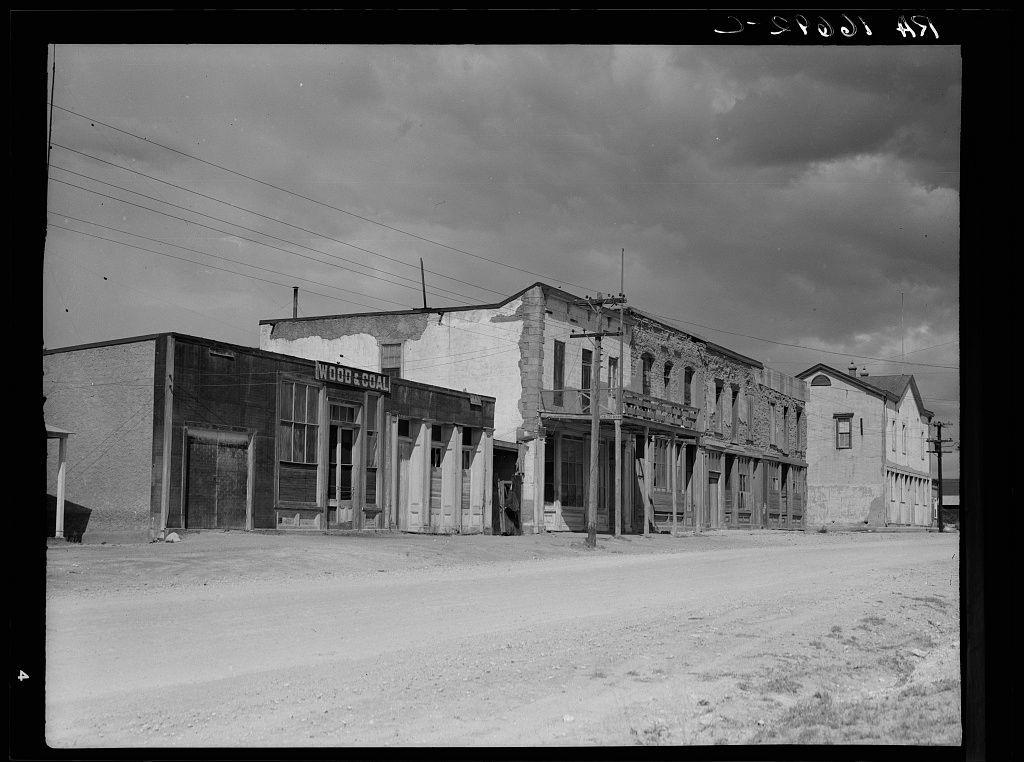 The black and white photograph depicts an old western-style street with several two-story buildings. The architecture features wooden facades, some of which have faded paint or graffiti on them. A prominent building in the center has a sign that reads "Wood & Coal," indicating it may be related to lumber goods during its operational period. This could suggest the photo is from an earlier era when such businesses were common.

The street appears deserted with no visible pedestrians, vehicles, or moving objects, which adds to the historical ambiance of the scene. The buildings show signs of wear and age, including peeling paint and boarded-up windows, suggesting they are not in use today. Overhead power lines stretch across the sky, a contrast between old architecture and modern infrastructure.

The image is labeled with "Dorothea Lange" on top right corner followed by an illegible number at bottom left indicating cataloging or inventory information likely used for archival purposes. Below that text reads "J-SPUD 42," potentially denoting the photographer, sequence in a series of photographs, and date.

The ground is unpaved with dirt roads typical of late 19th-century Western towns. The overall composition captures a snapshot of life from what appears to be an earlier period, possibly during or after the peak years of mining activity which was common in places like Tombsto [...]