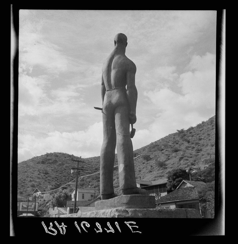 The image depicts a large bronze statue of a male figure standing on a pedestal in an outdoor setting. The sculpture is highly detailed, showing muscular definition and the texture of skin, with notable emphasis on the torso and arms. In one hand, it holds what appears to be a pickaxe or shovel handle, suggesting laborious work such as mining.

The background reveals a hilly landscape typical of arid regions, indicative of an open-air environment possibly within a desert area like Arizona. Buildings are visible in the distance, nestled among trees and shrubs that line the hillsides, which adds to the rural ambiance of the scene.

At the bottom right corner, there is handwritten text "Kit Carson" written vertically down through horizontal lines. In addition, at the lower left side, another inscription reads "Dorothea Lange," likely attributing the photograph or crediting its creator.

The image exhibits a monochromatic tone with high contrast between light and shadow areas, which accentuates the sculpture's form against the natural landscape backdrop.