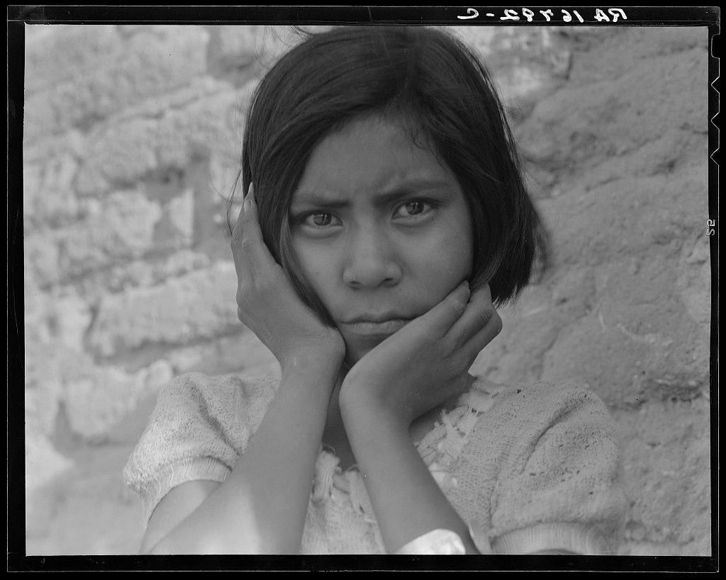 The black and white photograph captures a young girl with dark hair against the backdrop of a rough, brick wall. She appears pensive or possibly upset, her gaze directed off to the side rather than at the camera. Her hands are gently resting on either cheek, fingers spread out across her cheeks, suggesting she is deep in thought or concerned about something not visible within this frame.

The girl's clothing suggests it might be a casual setting: a white knitted sweater with some frayed edges around the collar and sleeves indicates wear that could come from laborious work. The texture of the wall behind her appears weathered and worn, further implying an outdoor environment possibly associated with rural or agricultural settings. 

There are handwritten characters at the top right corner which read "3-28 51," likely a date stamp indicating when this photograph was taken in March 1951.

The image is credited to Dorothea Lange and provides context as part of her collection, specifically linked with Mexican field laborers near Chandler, Arizona. This suggests the girl could be related to one or more such workers who were subjects for Lange's work documenting socio-economic conditions during that period in America.