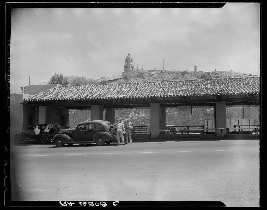 The image is a black and white photograph depicting an outdoor scene at what appears to be an inspection station located on the California-Arizona state line. It features a large building with tiled roofing, indicative of Mediterranean or Spanish colonial architectural styles commonly found in southwestern regions like Arizona.

A vintage automobile parked alongside the curb has its hood open as two individuals engage in conversation beside it. These people are likely inspectors and travelers at this agricultural inspection station to prevent the spread of plant pests across state lines.

In the background, a distinctive bell tower with multiple bells can be seen atop a hill, which may belong to a church or other significant building overlooking Yuma, Arizona. The sky is partly cloudy but allows ample sunlight illuminating the scene.

The image's caption references its historical context as part of California Department Agriculture efforts in 1938 during times when agricultural inspections were vital for controlling pests and diseases that could affect crops across state lines.