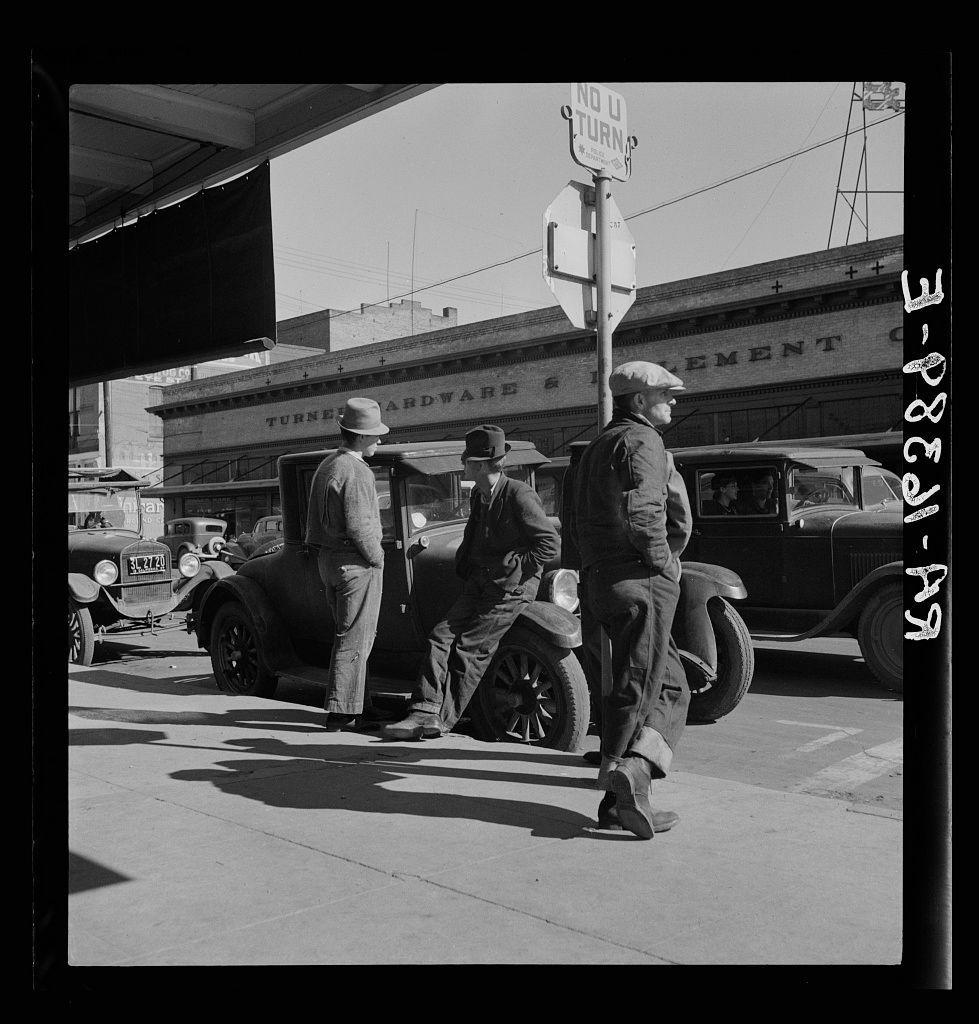 The black and white photograph captures a moment from the past on an urban street corner, where men are gathered around vintage vehicles. One man stands near a stop sign that reads "No U Turn," with his back to us, while another leans against what appears to be a turnstile or meter in front of him. To their right is an individual leaning casually onto a classic car's open door. The setting suggests this may have been taken during the Great Depression era given the worn-out appearance of both people and vehicles present.

The scene depicts everyday life with individuals dressed in work attire, indicative of men possibly engaged in manual labor or related activities common to that time period. Their clothing is practical and suitable for outdoor work—oversized hats, coats, trousers, and sturdy boots are prominent features.

In the background, an industrial building bearing a sign "Turner's Hardware & Improvement" provides context about the setting being near commercial establishments. The overall composition of this photograph suggests it might have been taken in Modesto, California as indicated by the reference to Men on 'Skid Row.' This area was known for its gritty atmosphere during that period and is often associated with social issues such as poverty.

The image evokes a sense of historical realism, providing insights into societal conditions through visual documentati [...]