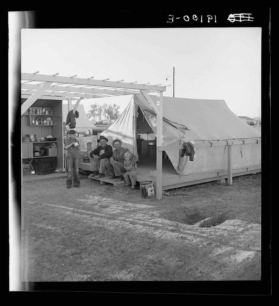 The black and white photograph depicts a temporary outdoor living situation. It appears to be at a migrant labor camp, specifically linked to the Farm Security Administration (FSA), located in Brawley, Imperial County, California. Four individuals are present: two adults seated on platforms outside of a tent-like structure with rolled-up sides for ventilation or sunlight exposure control; one adult standing inside another sheltered area within the makeshift setup, and a child perched on an elevated platform beside them.

The environment is utilitarian, suggesting limited amenities but essential living quarters. The ground shows patches of dirt and sparse grass in what appears to be a semi-arid region typical for agricultural work conditions like those described by Dorothea Lange's photography. A utility cupboard with various items stored within it can also be seen beside the makeshift structure.

The image conveys a sense of community among its occupants amidst their transient living situation, reflecting broader social issues and labor migration during that era as documented in FSA projects which aimed to capture human conditions across America through her lens.