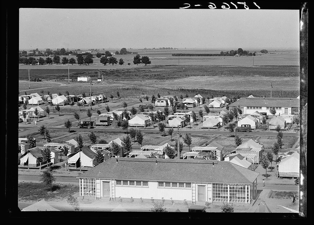 The image depicts a large outdoor settlement with numerous white structures that resemble temporary housing units. These are arranged in rows and spread across an expanse of land, suggesting the area is likely used for accommodating people on a short-term basis.

Each structure appears to be similar but not identical; they have pitched roofs and various types of windows or openings, some covered by what looks like canvas material. The settlement has trees planted at intervals along pathways, adding spots of greenery amidst an otherwise barren landscape.

The background features open fields with patches of crops in the distance, indicating that this area is agricultural land. A few larger buildings are visible as well; one large building appears to have a garage or workshop section and several rooms for habitation above it, possibly used by camp staff or supervisors.

In terms of vehicles, there's an old-fashioned truck seen driving through the settlement on what seems like a dirt road. The absence of modern amenities suggests that this might be from mid-20th-century America during a time when such camps were common in rural areas for people working in agricultural industries.

The sky is overcast with clouds diffusing sunlight, which gives an overall muted and neutral lighting to the scene. There's no visible activity or movement within these structures or on their surrou [...]