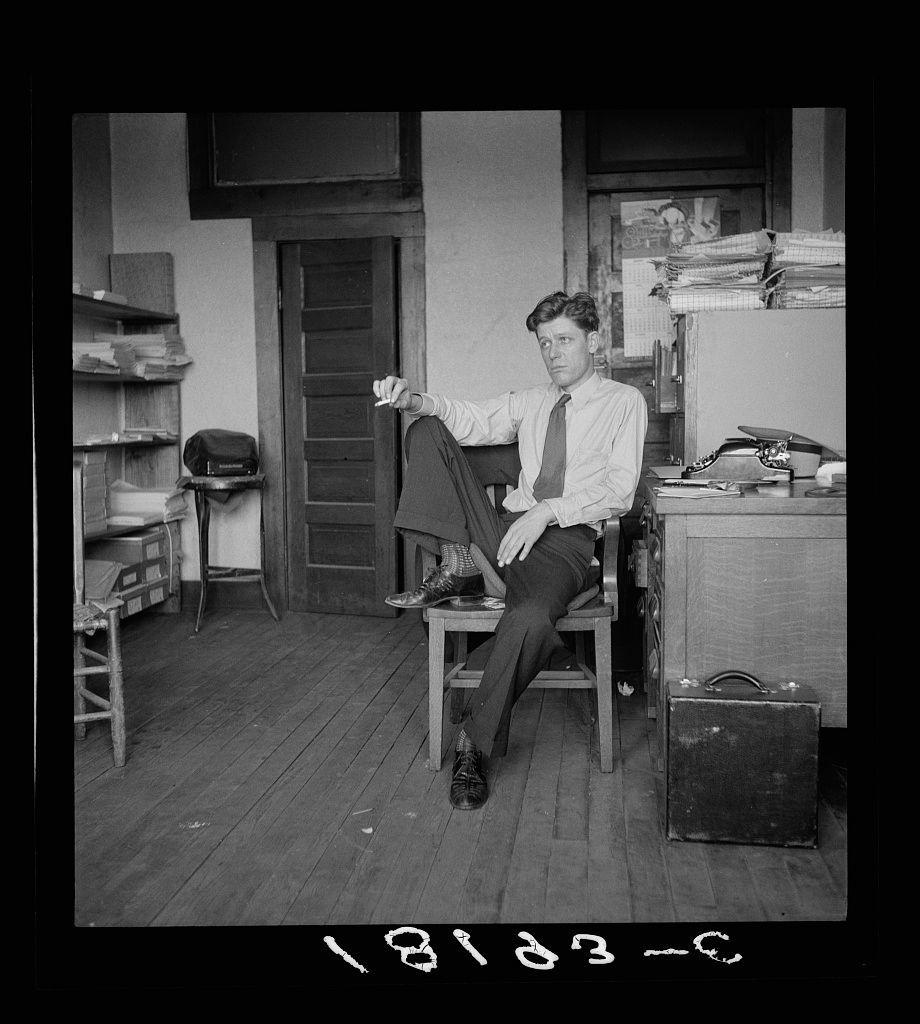 This black and white photograph depicts a young man seated in an office environment from the early 1930s, judging by his clothing style and hairstyle. He is dressed semi-formally with a light-colored dress shirt, dark trousers, and tie, paired with polished leather shoes that are not worn but rather placed on top of the chair beside him. His posture suggests casual confidence as he rests one leg atop the other while holding what appears to be a pencil or pen in his hand.

The office is modestly furnished with various items scattered around: an open suitcase at his feet, shelves and drawers filled with papers and books against the wall behind him, stacks of documents on top of another piece of furniture beside him. A filing cabinet sits nearby, partially obscured by these piles. The room's door stands ajar in the background.

The image is marked "1930-2" at its bottom right corner, indicating it was taken during that year or possibly referencing two years since 1830, which could be part of a larger numbering system not fully visible here. On the top left side, there's faintly discernible text likely related to documentation details about this photograph.

The overall ambiance suggests an individual who is either taking a momentary respite from work or posing for a candid portrait capturing his daily life in that period.