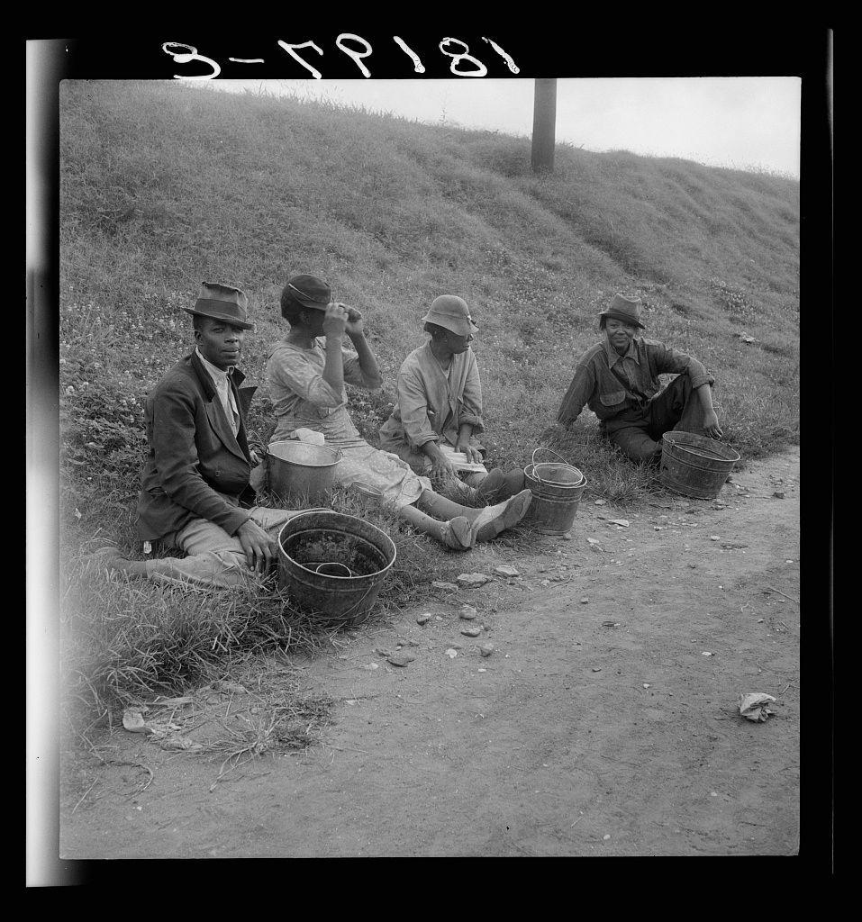 A black and white photograph depicting four individuals seated on a dirt path beside a hillside. They appear to be taking a break from work, as suggested by the buckets placed nearby containing what could be berries or other produce they've collected. The group is dressed in casual outdoor clothing suitable for manual labor; some wear hats while others don coats and suits, indicating varied levels of formality despite their shared activity. One person is using binoculars to look over a distant horizon, suggesting contemplation or supervision. There's a handwritten inscription at the top stating "3-81," potentially denoting March 1945 as it appears in Roman numerals (MMLXLV). The location mentioned provides insight into their profession and economic status—Berry pickers awaiting transportation to work for an estimated earnings of $0.60 per day, in Memphis, Tennessee during a historical period marked by labor strife or social changes.