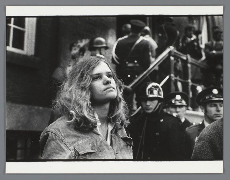 The image is a black and white photograph featuring an individual with long, wavy hair gazing off to the side. The person appears contemplative or possibly concerned as they look away from the camera. They are wearing what seems to be a denim jacket over casual clothing.

In the background, there's a group of people who appear to include both civilians and police officers. One officer is prominently visible in uniform with a helmet that has a reflective visor, indicative of law enforcement presence during an event or situation requiring their attention. The setting appears to be outdoors near some stairs leading into a building.

The overall atmosphere suggests a public gathering or protest scene from the late 20th century, judging by the fashion and photographic style. There is no explicit indication of specific events beyond this interpretation.