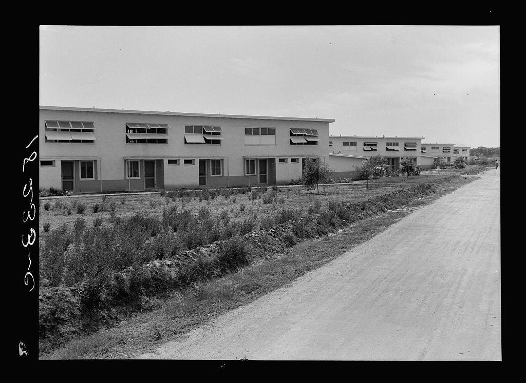 The image displays a black and white photograph of what appears to be an apartment complex. It features several identical two-story buildings aligned in parallel rows, with multiple rectangular windows on the façade indicating individual living units or apartments within each building.

The architecture is simple, functionalist design typical for housing projects during mid-20th century America; flat roofs and a repetitive layout are evident. The exterior looks utilitarian without much decoration, focusing more on utility than aesthetics. There's an expansive dirt road in the foreground that stretches towards one of these buildings, suggesting easy access to this part-time residence.

The environment is quite barren with overgrown weeds around what appears as undeveloped land behind and beside the housing units, indicating a rural or underdeveloped area rather than urban density. The sky above is clear without any visible clouds which implies it might be midday given that shadows cast are relatively short. No people can be seen in this photograph.

There's handwritten text on the edge of the image with what seems like numbers and letters scribbled but not entirely legible, possibly indicating a date or some other form of annotation relevant to the photo’s provenance or context within a collection such as FSA (Farm Security Administration).