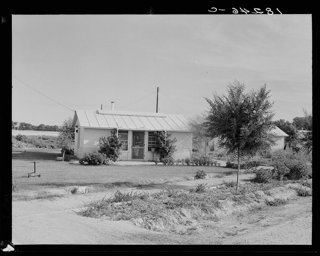 The image is a black and white photograph depicting a rural setting with an old, modest house as the central focus. The structure appears to be a single-story building with a gabled roof featuring two prominent horizontal ridges on its facade. There's a noticeable window covered by curtains at what seems like ground level, possibly indicating it could serve as a door or garage entrance.

The home has white siding and trim work that contrasts the dark windowsills and shutters. A wooden fence partially encloses the property in front of the house but is not clearly visible due to perspective. To the right side of the photo stands an outbuilding with what appears to be a corrugated metal roof, suggesting it could serve as storage or another utility space.

Surrounding the residence are various vegetation and shrubbery that adds texture to the scene, indicative of rural life but not well maintained, perhaps reflecting economic conditions. There's no visible sign of modern amenities like satellite dishes or contemporary appliances outside which might suggest a more affluent setting today.

A single tree with sparse leaves is notable in front of the house on its right side, possibly offering some shade and privacy while standing as a lone sentinel against the open field that extends into the distance beyond. The sky above appears clear, suggesting fair weather conditions at the  [...]