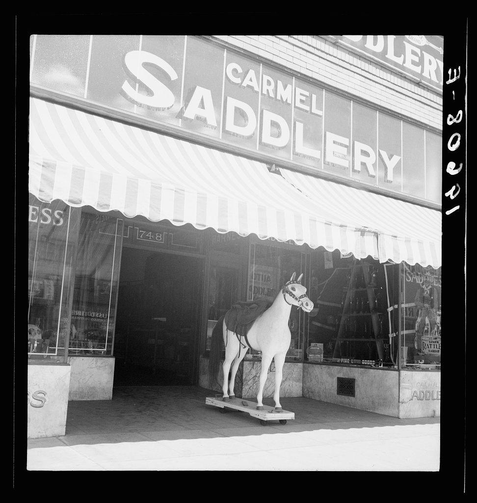 The image is a black and white photograph depicting the storefront of "Carmel Saddlery," which features signage above its entrance. A prominent feature in front of this establishment is a large-scale, life-sized statue or model of a donkey standing on what appears to be a small platform or board within the store's window display area.

The setting suggests it could be an older photo as indicated by the style and composition, with details like striped awnings over the storefront. The image carries a sense of historical context due to its monochromatic nature and subject matter.

Additional information about this particular photograph is sourced from Fresno on U.S. 99, indicating that the location may have some cultural or documentary significance related to Dorothea Lange's work. Further details can be found at https://images.loener.nl/DorotheaLange/full/6495/6495b7ceba25398d90c4f3f3.jpg.

This image captures the essence of a town in California, possibly from an earlier period when such saddlery stores were more common. It offers insight into local commerce and community life during that time through its representation of a specific store's character and storefront aesthetics.