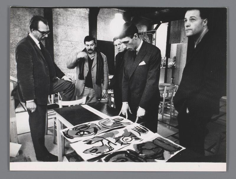 The image is a black and white photograph showing four men gathered around an art piece displayed on a table. The man standing to the left, wearing glasses and dressed in a dark suit, appears to be gesturing towards or discussing something with another seated man beside him who wears a striped blazer over a light shirt and has his legs crossed. Behind them stands two other men; one is looking down at what seems like an artwork on paper featuring abstract figures while the other gazes intently ahead.

The setting suggests an informal, possibly creative environment such as an art studio or gallery. The table displays multiple pieces of work with varying degrees of abstraction and expressionistic brushwork. There are chairs around a dining area in the background, indicating that this may be part of a larger gathering or event related to art appreciation or critique.

The photograph has a historical feel, likely dating from mid-20th century based on fashion and photographic style, which adds context for understanding its significance as mentioned in the accompanying text.