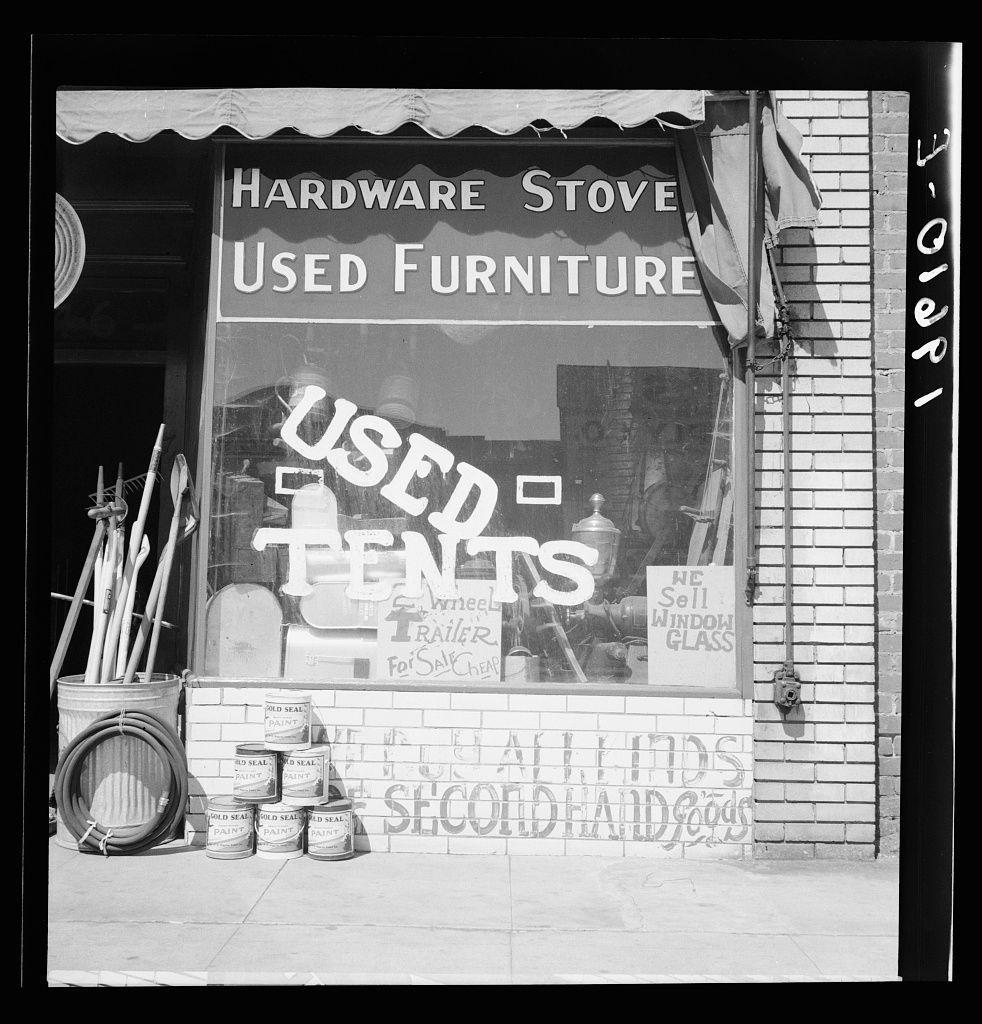 This black and white photograph captures a storefront with various items displayed outside, including what appears to be a hardware stove on the left side of the frame. The window displays signs advertising "Used Tents" among other items such as wheelbarrows, trailers for sale, and old seal paint cans stacked in three neat rows labeled "OLD SEAL PAINT." Above these signs is another that reads "We Sell Window Glass," suggesting a range of secondhand goods offered by the store. The storefront itself has signage indicating it sells "Hardware Stove" along with used furniture. The style of writing, as well as the overall image quality and composition, suggest an older time period.