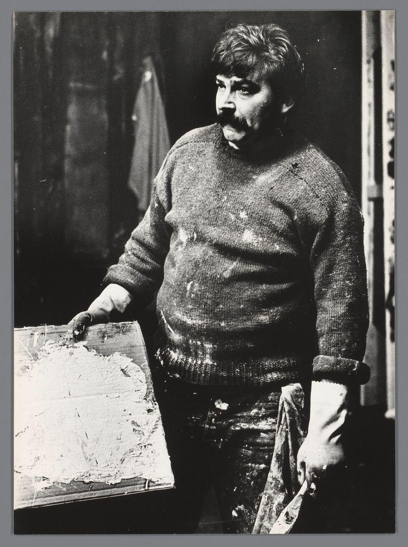 The image depicts a man standing with his right hand resting on an unfinished canvas. He appears to be in the midst of painting, as evidenced by paint splatters visible on his sweater and pants, along with brushstrokes still evident on the canvas. The artist has a mustache and is wearing glasses pushed up onto his head, suggesting he might have been working for some time without them due to discomfort or convenience.

He holds another canvas in his left hand, which looks like it's either being prepared for painting or recently completed but not yet cleaned up. His expression is contemplative, with a slight frown and intense gaze directed off-camera. The background suggests an indoor studio setting, dimly lit and filled with various art supplies and possibly more canvases.

The black-and-white photograph captures the artist in what seems to be a candid moment of deep concentration or reflection on his work. His clothing is casual yet functional for artistic pursuits, fitting comfortably against him despite being stained from painting activities.