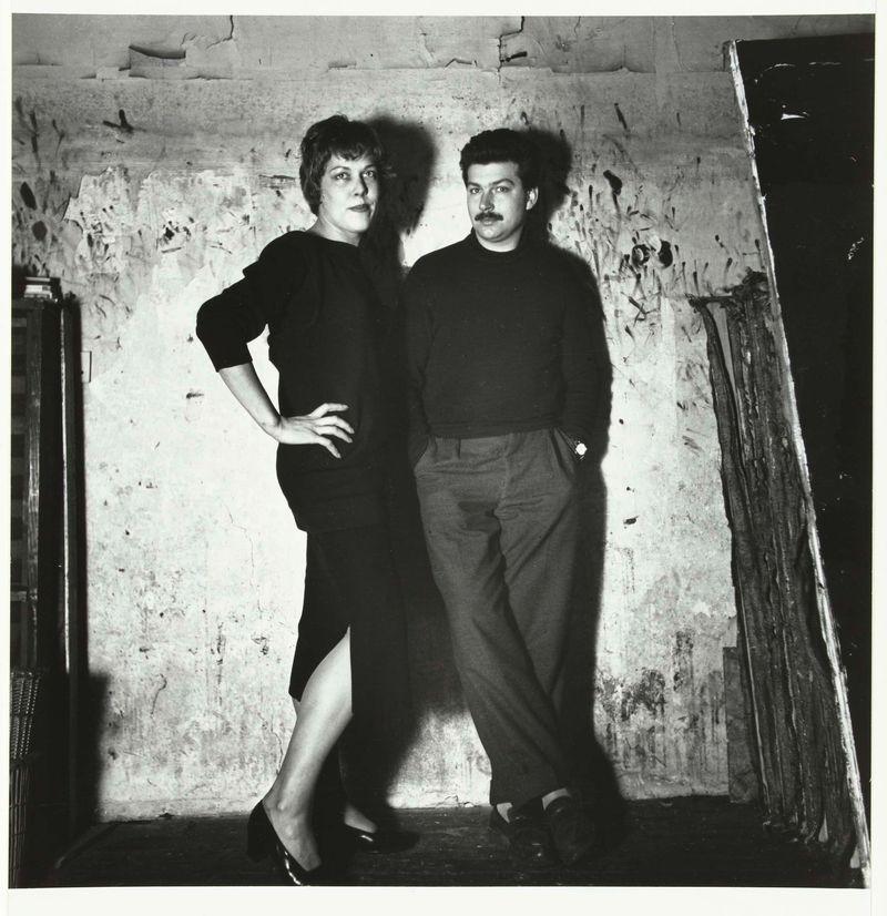 The image is a black and white photograph featuring two individuals, presumably Karel Appel and his wife, standing side by side against an unpainted wall with some visible texture. The person on the left appears to be wearing dark clothing, possibly a dress or top with short sleeves, paired with high-heeled shoes. They have their hands placed confidently on their hips, one hand resting on what looks like a metal object, perhaps part of the studio's equipment.

The individual on the right is dressed in a long-sleeved black shirt and trousers, standing with arms relaxed by their sides. The person has distinctive facial hair—a mustache—and appears to be wearing dark shoes. Both subjects have short hairstyles typical of earlier decades and are looking directly at the camera with neutral expressions.

In the background, on the left side, there's a glimpse of what could be studio equipment or storage items leaning against the wall. To the right edge of the photograph is an open door or frame leading to another darkened space, adding depth to the composition but not much detail about its contents can be discerned.

The lighting in this photo creates strong contrasts between light and shadow on their figures and the background, highlighting the textures of both subjects' clothing and skin. The overall mood conveyed by the photograph is contemplative yet poised, capturing a moment [...]