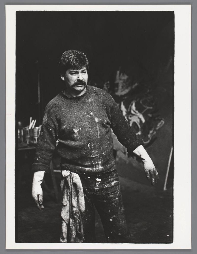 The image is a black and white photograph depicting an individual who appears to be in the midst of artistic activity, likely painting or sculpting. The person has dark hair with bangs that partially cover their forehead and wears a sleeveless garment smeared with paint. They are holding what seems like a cloth or piece of paper towel in one hand, while the other is free. Their facial expression conveys concentration or satisfaction.

The background reveals an environment indicative of a studio setting, filled with various items such as brushes or tools suggestive of art-making processes. The lighting highlights the individual and their immediate surroundings but leaves much of the space beyond into shadow, creating contrast that draws attention to them. There's no discernible movement captured in the still image.

The photograph is presented on a white background with what appears to be an edge of a page or frame at its top, suggesting it may have been printed from another source such as a magazine or album. The composition and lighting are typical for documentary photography capturing candid moments within artistic environments during that era.