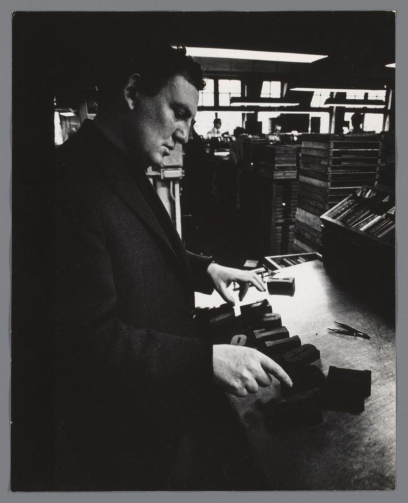 The image depicts a man working in an office environment with shelves of books or files behind him. He is dressed formally, wearing glasses and a dark suit jacket over what appears to be a dress shirt. The monochromatic tones suggest the photo was taken many years ago. His attention is focused on handling various rectangular objects which could be records or compact discs, indicative of work involving media archiving, possibly for music-related projects given his attire's formality suitable for high-profile events like 'Love Song'. No other people are visible in this frame. The image has a documentary feel and references Hugo Claus' collaboration with Ed van der Elsken on the creation of what is claimed to be the largest book ever made about him, titled "Love Song".