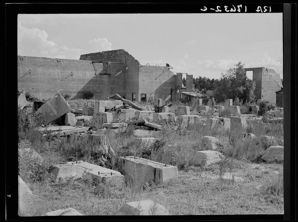 The image depicts a scene of desolation featuring remnants of what appears to be an industrial or construction site. The structures are in various states of disrepair, with broken walls and scattered debris such as concrete blocks and wooden planks strewn across the ground. Overgrown vegetation is visible throughout the area, indicating that nature has partially reclaimed the space after abandonment.

The sky above is clear with a few clouds, suggesting an open outdoor environment. The photo captures a wide view of the site, showcasing both vertical structures and flat surfaces filled with broken materials. There are no people or animals present in the scene, emphasizing isolation and decay.

This image seems to be part of historical documentation, potentially linked to abandoned industrial areas like Fullerton, Louisiana as mentioned in the provided caption. The black-and-white format gives it a timeless quality, which could indicate that these structures were once operational but have since been left to deteriorate over time.