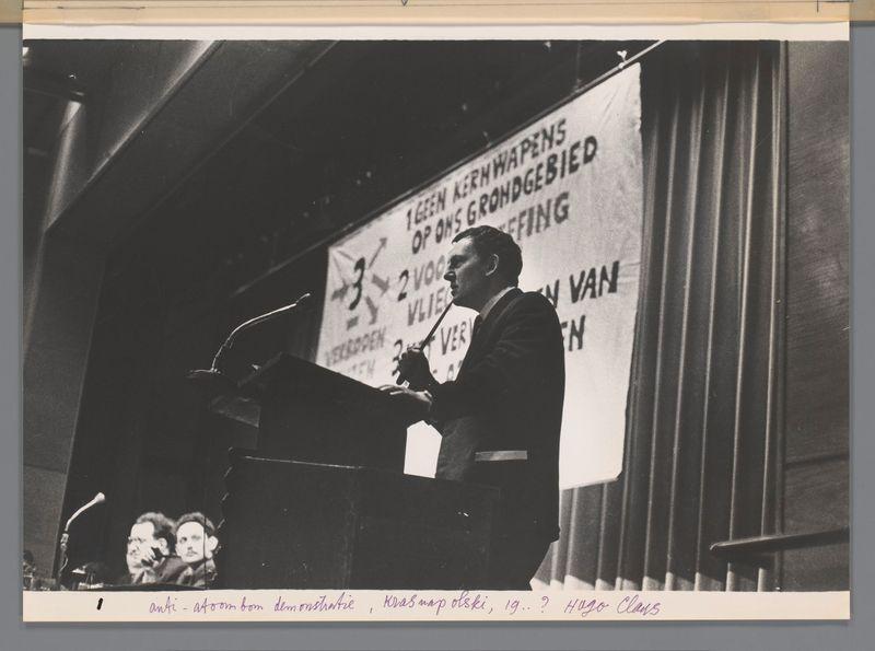 The image is a black and white photograph depicting an indoor scene with several individuals. In the foreground, there's a man standing at a podium speaking into microphones, likely addressing an audience out of frame. He appears focused on his speech or presentation, holding what seems to be notes in one hand while gesturing lightly with the other.

Behind him is a large banner containing text that suggests it may relate to youth activities involving groups and assemblies ("TEEN AGENDA KERKWAPENS VAN OPZICHTIGES"). The visible part of the word "VER" indicates some form of activity or assembly. There's also an indication of two voices, as suggested by one phrase on the banner.

In front of the podium are seated individuals; their expressions and postures suggest attentiveness to the speaker at the podium. One person appears to be taking a photograph with a camera placed between them.

The overall setting looks like a formal or semi-formal event, possibly related to youth activism given hints from the text on the banner. The photo has handwritten annotations in purple ink across the bottom and corner of the image that seem to indicate context such as "anti - atomic bomb" reference at Krasnapolsky Hotel Amsterdam by Hugo Claus (1961).

The style is indicative of mid-20th-century photography, with a clear focus on the speaker and audience interactions.