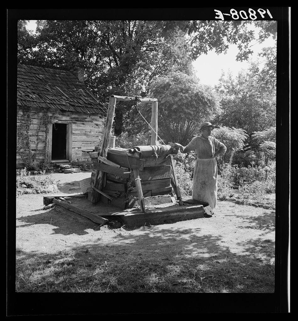 The image depicts a historical scene featuring an individual operating what appears to be an old-fashioned well pump or water drawing machine. This person, likely female based on the clothing and physical build, is standing beside the apparatus which has wooden components with metal fittings attached at various angles, suggesting it's used for manual labor in obtaining liquid from underground.

The setting includes a rustic environment indicative of rural life perhaps during early to mid-20th century America. A decrepit structure made of weathered wood can be seen behind and partially obscured by the subject, which seems out-of-use or abandoned with its broken window panes and overgrown surroundings suggesting neglect or disuse.

The well pump apparatus is positioned on a makeshift platform constructed from planks lying flat on the ground, showing signs of wear. The backdrop features lush greenery likely consisting of trees indicative of rural woodland areas common in historical American landscapes.

The photograph itself has visible text at its top edge: "3-808PI", which could be an identifier for cataloging purposes or a part reference number associated with this image within the context it belongs to. The black and white format enhances contrasts, emphasizing textures such as wood grain and foliage density against sunlight filtering through leaves.

The overall atmosph [...]