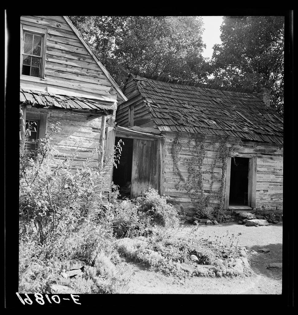 The photograph captures a dilapidated wooden structure, likely an abandoned house or barn. It is presented in black and white which gives it an aged appearance, consistent with historical photographs from the early 19th century.

From this vantage point, we see part of what seems to be two buildings connected side by side; they are both made of weathered wooden planks that have visibly deteriorated over time. The roof appears old and is composed primarily of shingles which look worn out or broken in places. 

The house's entrance is open with its door ajar, suggesting it has not been used for quite some time as there seems to be no activity around.

In the foreground lies an untended yard filled with weeds and overgrowth that further emphasizes abandonment. The vegetation varies from shrubs to taller plants indicative of wild growth in places often neglected by human maintenance.

There is visible damage on the structure, such as exposed beams and loose planks, hinting at years without repairs or upkeep. This image evokes a sense of desolation but also stands as a testament to resilience; it has survived through times where perhaps no one else had bothered with its care. The overall atmosphere suggests that this place was once inhabited and lived in with some degree of regularity before falling into disuse.

A unique identifier '13810-E' is stamped on the image, which cou [...]