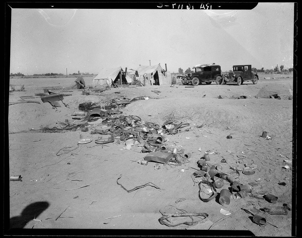 The image is a black and white photograph depicting a desolate landscape with scattered debris on the ground. In the background, there are makeshift tents or shacks lined up in an orderly fashion, suggesting some form of temporary settlement or campsite. Various discarded items such as metal containers, utensils, pipes, and other refuse litter the area around these structures.

In front of the huts, two older-style automobiles are parked on what appears to be a dirt road or clearing. The vehicles look dilapidated with visible rust and wear marks, indicating they have been exposed to harsh conditions over time. One car is closer to the foreground while another one is slightly further away from the viewpoint.

The sky above is clear without any clouds in sight, hinting at dry weather conditions typical of a desert or arid region. The overall atmosphere conveyed by the image evokes feelings of abandonment and hardship faced by people living within this setting.