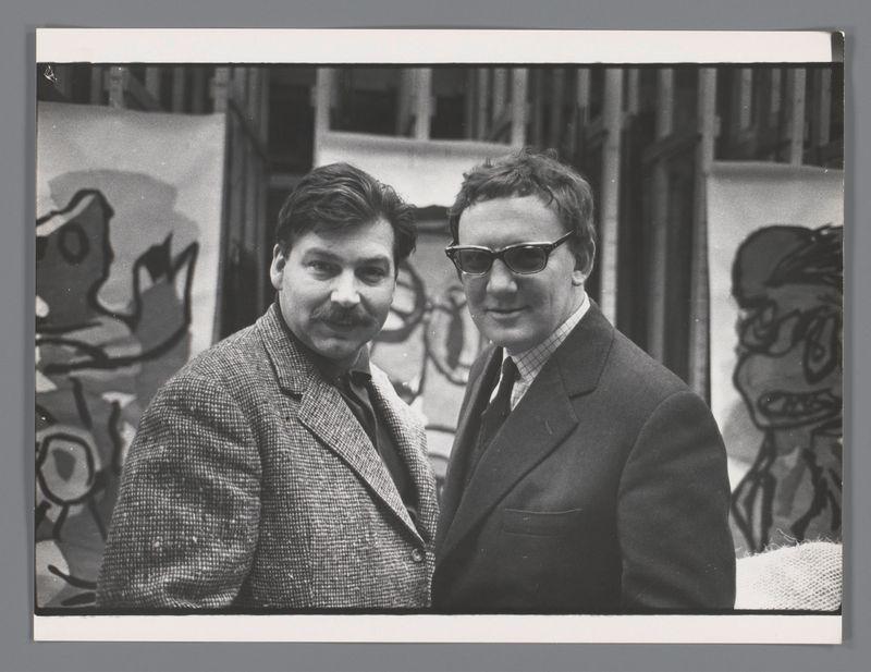 The image is a black and white photograph featuring two men standing side by side, posing for the camera. The man on the left sports a mustache, wears glasses, and has curly hair. He dons a patterned tweed jacket over a plaid shirt paired with a dark tie. His expression appears friendly as he gazes directly into the lens.

The individual beside him is dressed in a suit with an open collar showing a striped shirt underneath. This person also sports sunglasses resting on their head and has his hair parted to one side, giving off a more formal appearance compared to his companion's casual style.

In the background, there are abstract graffiti artworks visible, suggesting that this photo might have been taken at an art event or exhibition space. The artwork includes various shapes and lines with no discernible figures but hints of form in some areas which could be interpreted as human-like silhouettes or stylized characters.

The overall setting gives off a creative atmosphere, potentially connected to the arts scene mentioned in the additional information provided about this image.