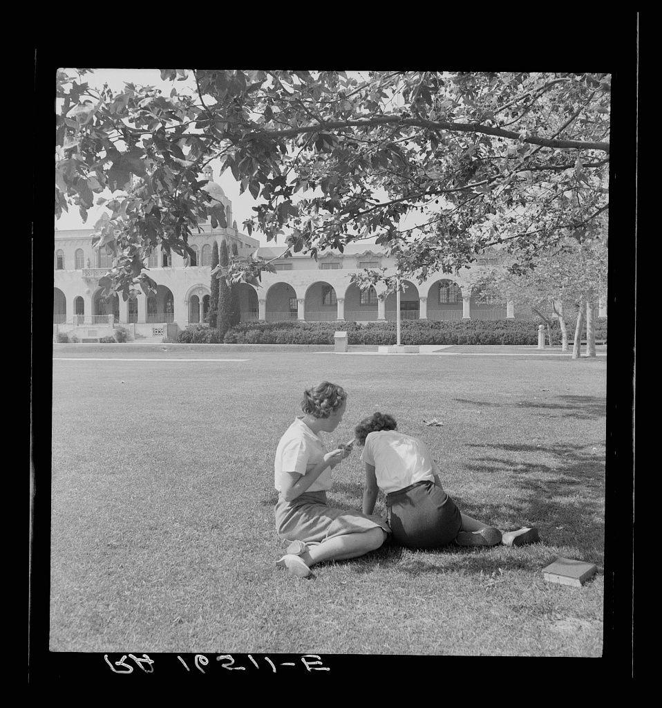 The image is a black and white photograph depicting two women sitting on the grass in what appears to be an expansive outdoor area. They are focused on reading or writing something, with one woman seated comfortably while holding papers, and the other bent over as if examining something closely.

In the background stands a grand building featuring multiple arches and domed structures, reminiscent of Spanish Colonial Revival architecture commonly found in California during that era. The grass is well-maintained, and there are trees providing shade around the area where the women sit.

At the bottom left corner of the image, text indicates "LS 192-46," which could be a reference to cataloging or identification numbers for archival purposes. This photograph seems to capture an everyday moment from mid-century America, possibly during a time when such settings were common places for leisure activities and social gatherings.

The additional information provided suggests that this image might have been taken at Fullerton High School in California as part of the Federal Art Project (FAP), which was established by President Franklin D. Roosevelt's New Deal programs to employ artists across America from 1935-43, primarily during the Great Depression era.

The photograph carries a sense of tranquility and simplicity, capturing an intimate moment between two individuals in a serene  [...]