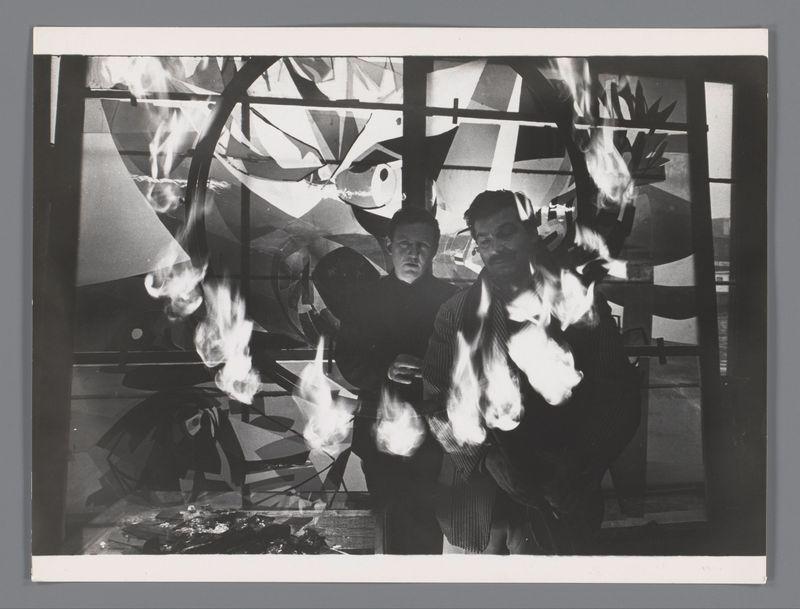 The image depicts a black and white photograph of two men in an artistic setting. The background consists of large, abstract shapes that appear to be part of a larger installation or painting with dominant shades of grey and black. Flames are prominently featured around the figures; several tongues of fire reach upward from what seems like burning objects on the ground.

The man on the left is wearing dark clothing and has his gaze directed towards something outside the frame, while the other person, dressed in lighter colors, looks directly at the camera with a contemplative expression. The intensity created by the flames adds dramatic effect to their expressions.

This photograph likely captures Hugo Claus and Karel Appel during the creation of "Love Song," commissioned for Andreas Landshoff's opus magnum 't Grootste Boek ter Wereld', as per external information provided. It conveys a sense of creativity, urgency, or intensity often associated with significant artistic endeavors.

Additional context indicates that this image is part of Ed van der Elsken’s work and holds historical value in documenting the early 1960s art scene involving prominent artists like Claus and Appel.