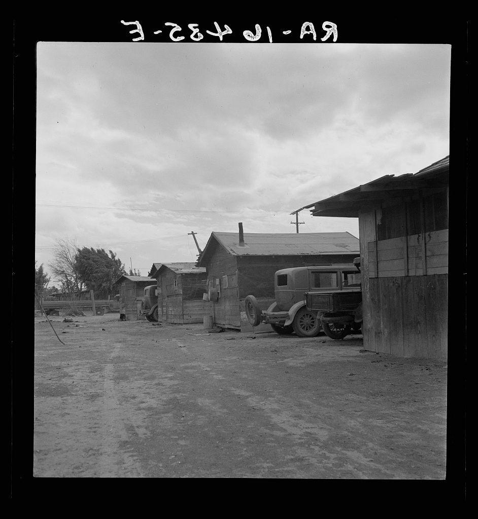 The image depicts an outdoor scene from what appears to be the early 20th century. It shows a barren, dirt-covered area with several dilapidated wooden structures and sheds of varying sizes. Some appear to function as makeshift homes or storage units.

In front of these buildings is parked a vintage vehicle resembling a Model T Ford, indicative of its age. The ground looks uneven and unpaved, suggesting it's an older rural setting without modern infrastructure. Power lines are visible in the background with one pole extending higher into the sky than others.

The image appears to be black and white or sepia-toned, giving it a historical feel. Overlaid on top is handwritten text that could represent metadata such as date or description related to archival information about slums of East El Centro, California where Mexican field workers lived in housing rented for eight dollars per month.
