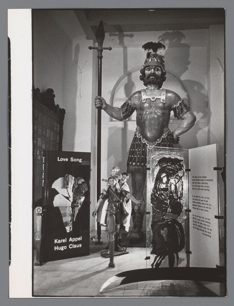 The image depicts a large statue of a muscular figure wearing classical armor, holding a spear and standing atop another sculpture that appears to be in fragments. The larger statue has exaggerated features such as an oversized head with facial hair and prominent ears. It is adorned with what looks like a crown or helmet on its upper part. Behind the statues are two displays: one reads "Love Song" next to artwork, possibly indicating it's related to the book mentioned in the caption; another display mentions artists' names Karel Appel and Hugo Claus associated with this exhibit. The overall scene suggests an art gallery setting where classical themes interact with modern artistic elements.