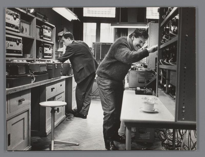 The image is a black and white photograph depicting two individuals working in what appears to be an electronics or laboratory setting. One person, wearing a sweater and trousers with dark shoes, leans into a shelf filled with various electronic equipment such as audio amplifiers and mixers, while the other individual stands nearby operating one of these pieces of equipment.

The environment is cluttered with numerous electronic devices and cables, suggesting that this space may be used for sound engineering or experimental work. A small white cup containing what appears to be a coffee mug rests on top of an amplifier in front of one person. The floor has scattered wires and tools indicating active use and maintenance. In the foreground, there is a stool near which another electronic device lies unattended.

The photograph captures a candid moment that conveys concentration and engagement with the technical tasks at hand. It appears to be from Karel Appel's time in the Studio for Electronic Music of the University of Utrecht during 1955-63, as indicated by the source information provided.