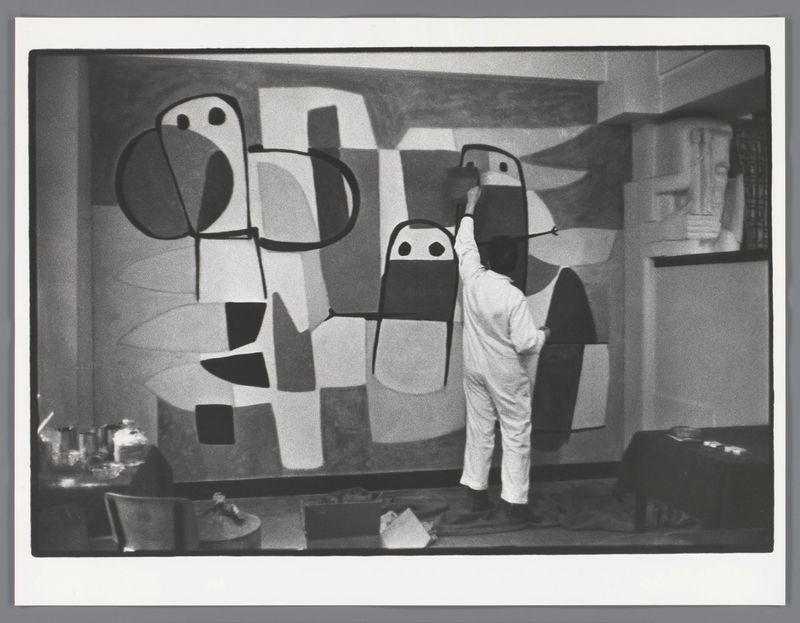 The image is a black and white photograph capturing an artist in the act of painting. The individual, wearing what appears to be all-white attire with rolled-up sleeves, stands beside a large canvas that features abstract forms composed primarily of geometric shapes such as rectangles, ovals, and circles filled or outlined with solid colors like black, grey, and shades of beige or cream. Some of these shapes have human-like faces drawn on them.

The artist is holding up what looks to be either a paintbrush or perhaps the edge of the canvas itself in front of one of his works-in-progress, possibly to examine it closely or consider how he might proceed with painting next. The studio setting includes various items scattered around such as furniture, containers, and other artistic paraphernalia, suggesting an environment focused on creativity.

The photograph is credited to Karel Appel during the restoration of his mural 'Vragende Kinderen' at the former town hall in Amsterdam, captured by Ed van der Elsken, circa 1959. The image's composition draws attention primarily to the artist and his interaction with the artwork on display, conveying a sense of immediacy and engagement within an artistic endeavor.