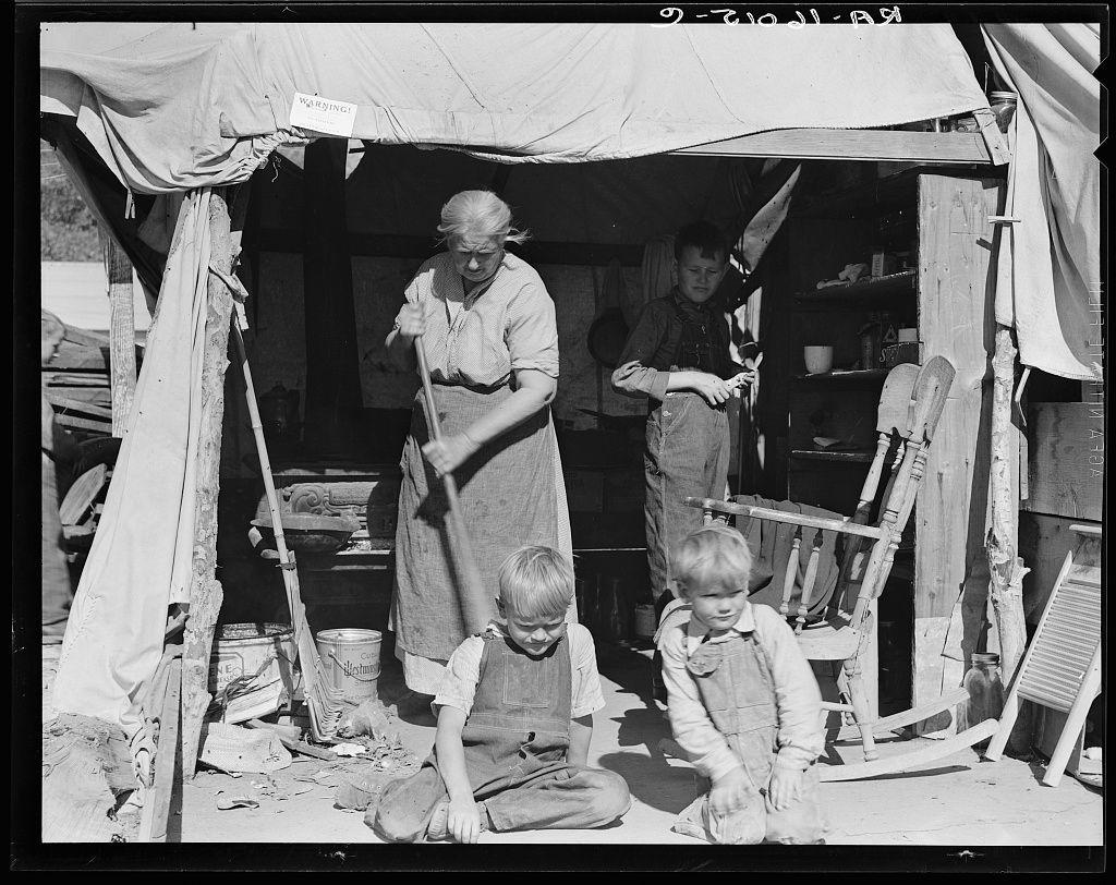 The image depicts a black and white scene of an aged woman standing in what appears to be a makeshift shelter or camp. She is wearing apron-like clothing, with one hand resting on her hip while the other holds up a broomstick vertically. To her right stands a young boy dressed in overalls, holding something small that seems like it could be a tool or toy. In front of him are two more children sitting close together; both also appear to wear similar attire and have their attention directed towards either the woman or another point off-camera.

The environment is cluttered with various objects: there's an overturned chair, broom bristles on the ground in disarray, a metal bucket labeled "Blackmore," and pieces of broken wood. The entrance features draped fabric tied back to reveal more items within. In the background, partially visible, are structures that suggest other dwellings or parts of camp life.

There is a sign with text above her head reading 'WARNING!' which could imply cautionary measures in place due to potentially unsafe conditions for children. Overall, this image conveys hardship and resilience amidst poverty or displacement, as indicated by the context provided from Aged woman with three of her twenty-two grandchildren, Kern county migrant camp, California.