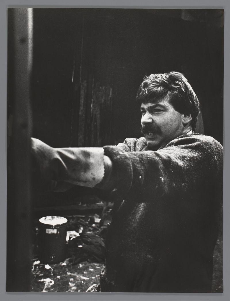 The image depicts a monochromatic photograph of an individual engaged in what appears to be a painting or sculpting activity. The person is captured with their right arm extended, holding onto something outside the frame while wearing a textured garment that suggests manual labor. In front of them on a surface lies a paint can and various other art supplies scattered about. The background features dark tones which provide contrast against the lighter-colored materials in use. Notable details include visible splatters or drips associated with painting, as well as what might be remnants of plaster or clay, hinting at an artistic workspace amidst creative chaos.