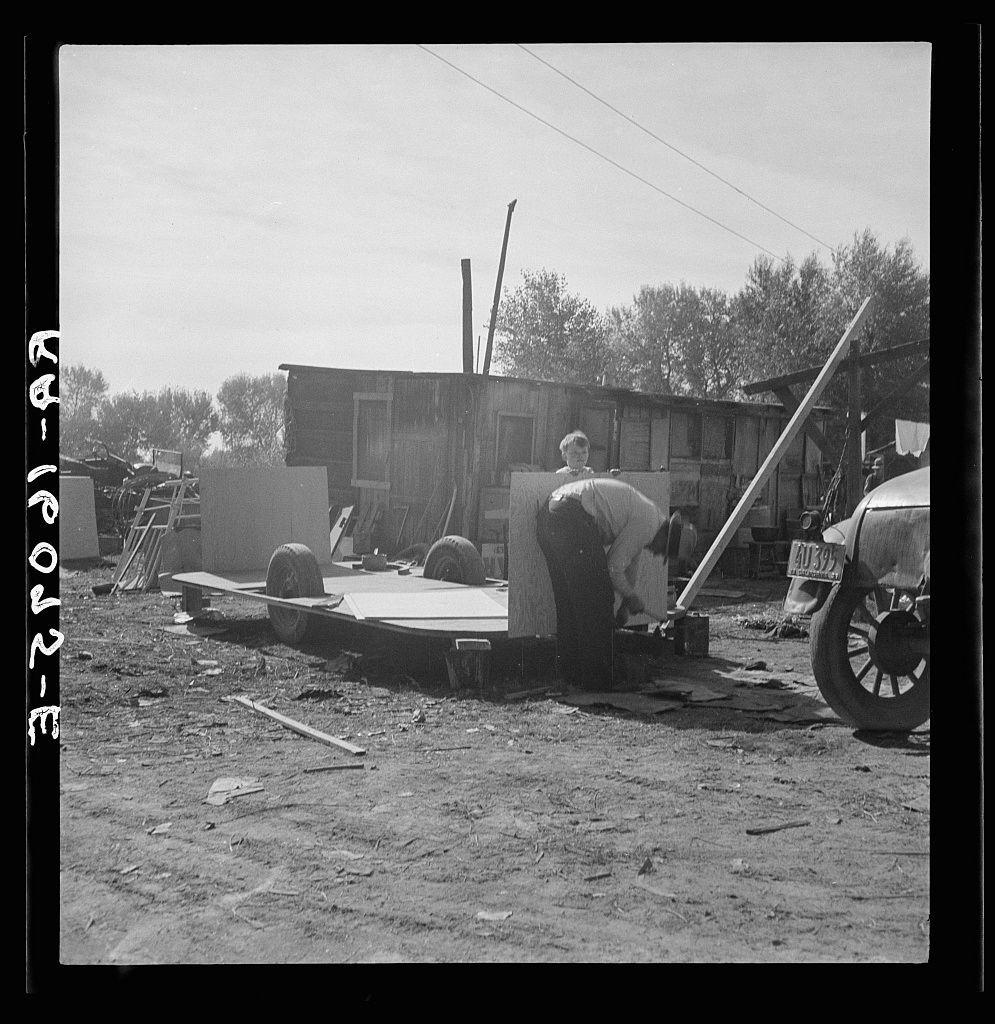 The image is a black and white photograph depicting an outdoor scene. In the foreground, there's a trailer with large wheels parked on what appears to be dirt or unpaved ground. A man in casual attire, including trousers and a long-sleeve shirt, is bending over near one end of this flatbed cart, seemingly engaged in some kind of work involving wooden panels placed horizontally across the bed.

To the right side, an older model car with visible rust marks on its body suggests it's been exposed to weather conditions for quite some time. The vehicle has a license plate attached at the front but not fully legible due to distance and angle. 

In the background stands what appears to be part of a makeshift structure or shack made from materials like wood, metal, and possibly old car parts, indicating this could be an improvised dwelling in a rural setting.

The environment seems somewhat desolate with scattered debris on the ground, including pieces of lumber and other construction-related waste. Overhead, power lines stretch across the sky, hinting at some level of infrastructure development nearby.

Trees are faintly visible behind what looks like a barrier or fence, suggesting this might be in an area where more greenery is possible but not immediately within sight due to ongoing work or clearance activities around.

The overall impression conveyed by the photograph suggests [...]