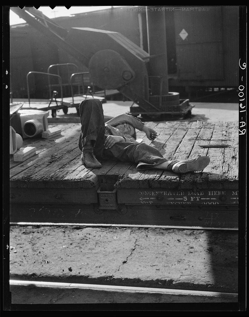 The image is a black and white photograph depicting an individual reclining on what appears to be the plank of a wooden dock or platform. The person, whose gender is not specified from this perspective, is dressed in work attire with cargo pants, boots, and casual shoes indicating potentially labor-intensive activities. One leg is bent at the knee while the other extends outward, suggesting a momentary rest during physical exertion.

The background features large industrial machinery indicative of heavy industry or shipyard operations; cranes are visible alongside a sizable machine that may be used for loading cargo onto ships, such as a dockside crane. The setting implies an outdoor environment where significant manual labor is performed, likely at a port or shipping facility in Sacramento, California.

The photograph carries historical connotations and evokes themes of toil, human resilience amidst industrial settings, possibly during the early 20th century given its black-and-white nature which suggests it's from that era. The presence of such machinery alongside human figures emphasizes the scale and gravity of labor within this context.