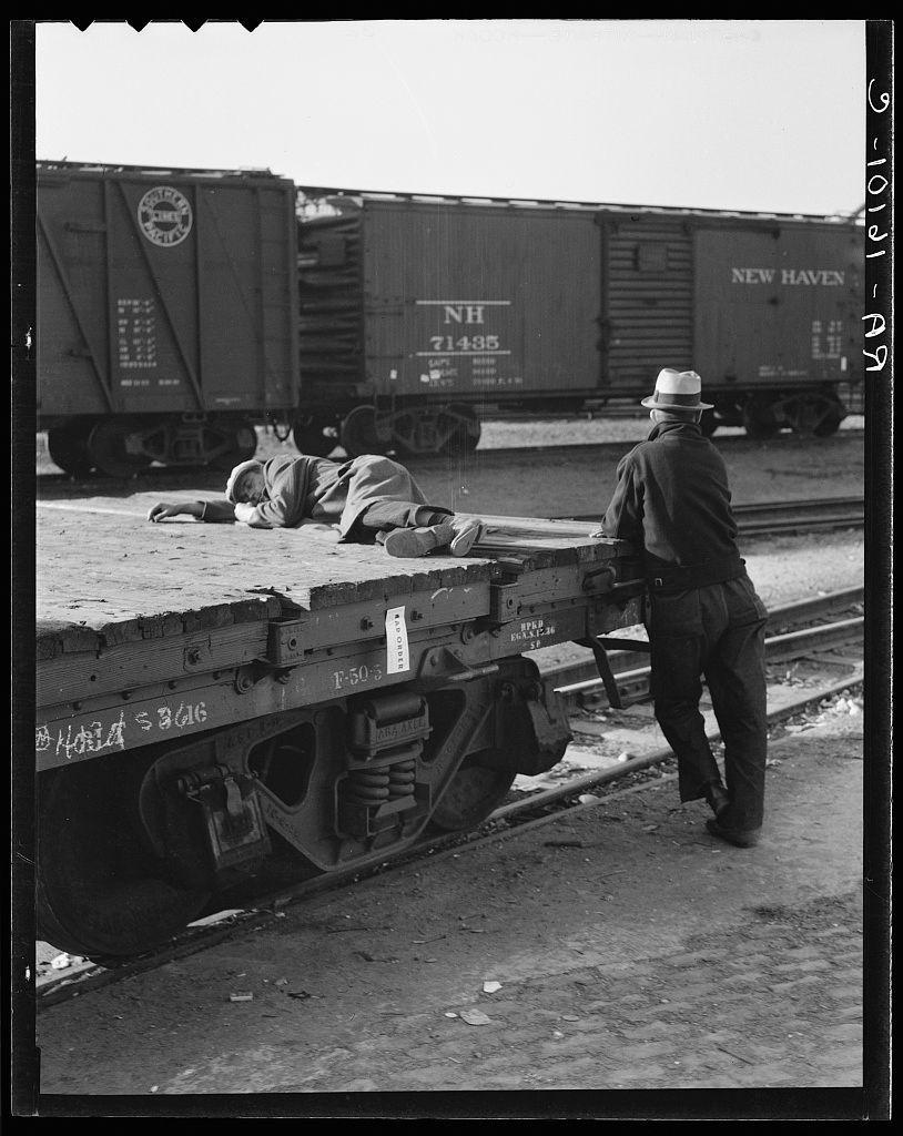 The black and white photograph depicts a scene within what appears to be a railroad yard. In the foreground, there is a railway flatbed or car with visible wheels labeled "Baldwin 19-6." A person wearing a dark coat, pants, shoes, gloves, and a hat stands on this railcar, leaning against it as if taking rest after laborious work.

In contrast, another individual lies face down in repose atop the flatbed. This resting figure appears to be dressed in casual clothing with an overcoat or similar garment draped across their body. The surroundings suggest a bustling industrial environment: multiple freight cars bearing "New Haven" and other inscriptions are stacked on tracks behind them.

The overall mood conveyed by this photograph hints at themes of exhaustion, labor, perhaps even despair within the context of manual work in America during an earlier period.