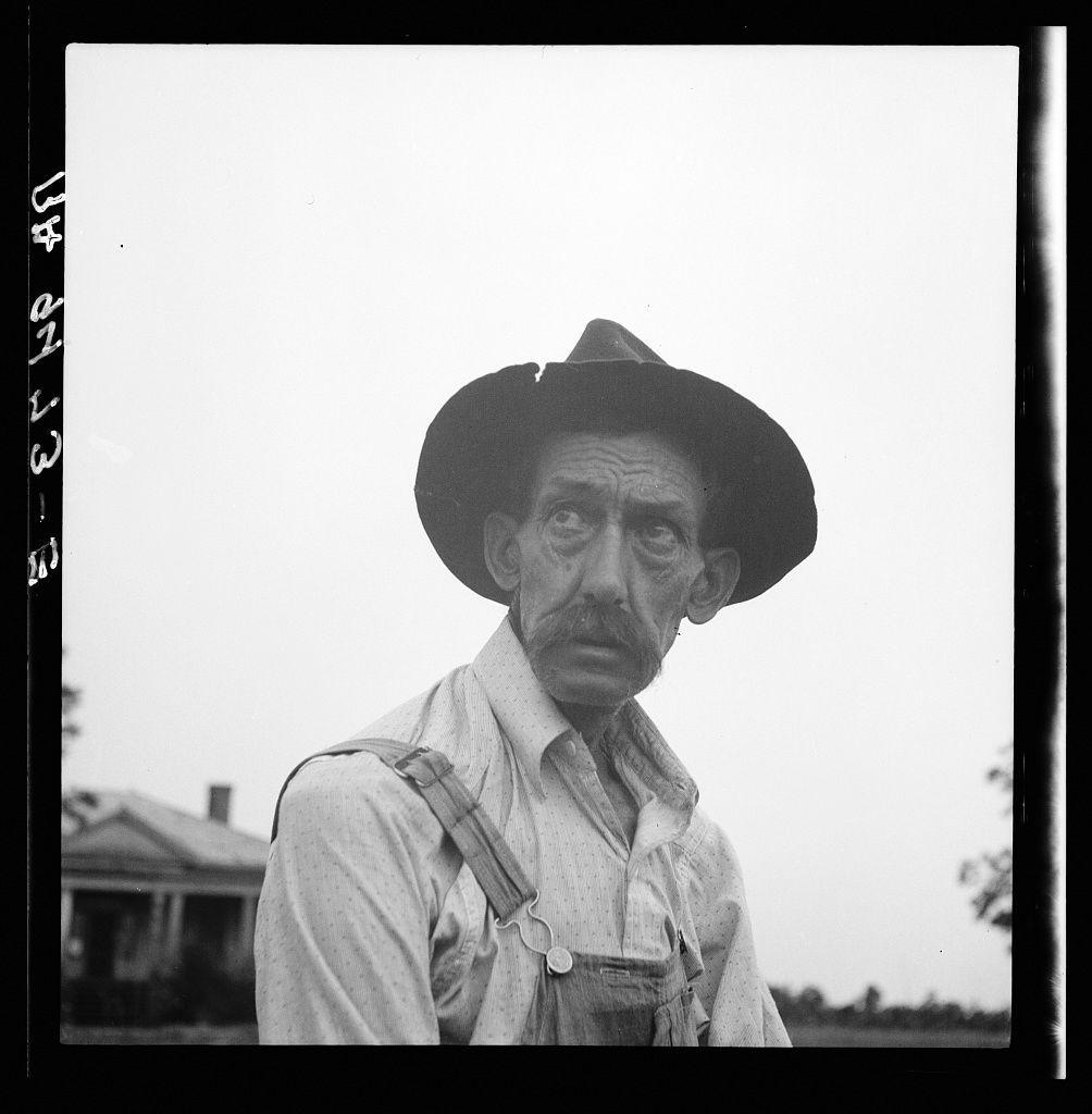 A black and white photograph shows an older man in the foreground. He wears a wide-brimmed hat, suspenders over his shirt, which has rolled-up sleeves, suggesting work attire suitable for farm labor. His expression is serious or contemplative as he gazes slightly off-camera to the right. In the background, there's a simple house with what appears to be a porch visible behind him. The sky above is overcast and featureless, indicating either early morning light or possibly an impending weather change.

The photograph bears markings on its borders which suggest it may have been part of a larger collection or album; these marks read "1934" in the top left corner and what appears to be a symbol resembling a film strip with various notations. The lower right edge shows a partial vertical mark, possibly from handling during storage.

The context provided indicates that this image is associated with Thomaston (vicinity), Georgia, relating it to Dorothea Lange's works documented in the Farm Security Administration collection for her project on rural life and American poverty in 1936. The man seems to be expressing concern or hardship regarding crop yield during a time of significant economic struggle, likely indicative of the Great Depression era.

This photograph is part of an ongoing narrative captured by Dorothea Lange through her lens, documenting human experiences within the  [...]