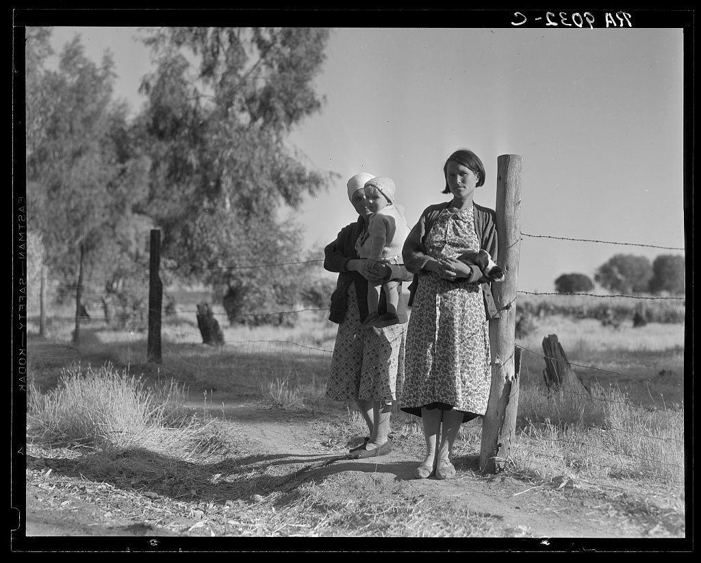 The image is a black and white photograph depicting two women outdoors, likely from the early to mid-20th century. The setting appears rural with dry grass on the ground and sparse trees in the background under what looks like clear skies.

On the left side of the photo stands a woman wearing a patterned skirt over pants, long sleeves, and closed-toe shoes. She is holding an infant wrapped snugly against her body; only the child's head protrudes from underneath a light-colored bonnet or hat. Her expression seems neutral as she gazes off to the side.

Beside her stands another woman wearing what appears to be a floral dress with short sleeves and ankle-length hem, complemented by closed-toe shoes. She is also holding an infant; however, this child's features are more visible due to not being wrapped in fabric. This second woman wears a wide-brimmed hat on her head.

A rustic wooden fence runs diagonally across the foreground of the photograph with barbed wire attached at its top section, indicating it might be enclosing farmland or property boundaries. The overall atmosphere suggests themes related to rural life and possibly migrant workers during a time when such conditions were prevalent in certain regions like California's agricultural areas.