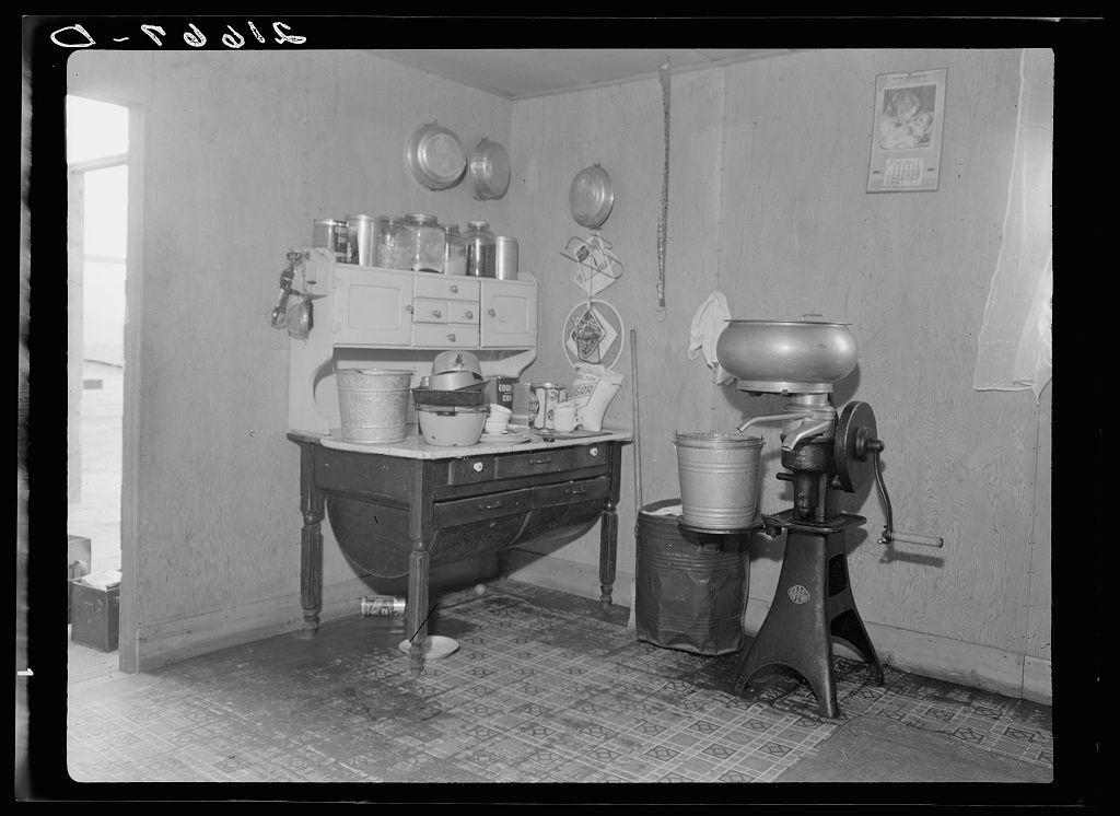 The image is a black and white photograph depicting the interior of an old-fashioned kitchen or pantry. It shows various cooking utensils, including canisters on shelves above a wooden table with drawers underneath it. On top of the shelf are several jars and tin cans organized neatly in rows.

To the right side, there's a manual grain grinder attached to a large base, which is connected by an extended handle for turning. Beside it, closer to the wall, stands a small bucket placed on what appears to be a wire mesh or strainer within another larger container that might serve as a trash can.

The floor has patterned tiles with multiple colors and shapes. On the back wall hangs some papers including a calendar, suggesting everyday use of this space for domestic activities such as cooking, preserving food items like jams or pickles, which was common in rural settings during earlier times. The overall setting gives an impression of simplicity, functionality, and preservation typical of agricultural lifestyles from past centuries.

Additional information suggests that the photograph belongs to Dorothea Lange (1895-1965), a pioneering photojournalist known for documenting various aspects of American life throughout her career spanning more than three decades. This image specifically is identified as belonging in 'Cut-over area', which refers possibly to an isolated community or  [...]