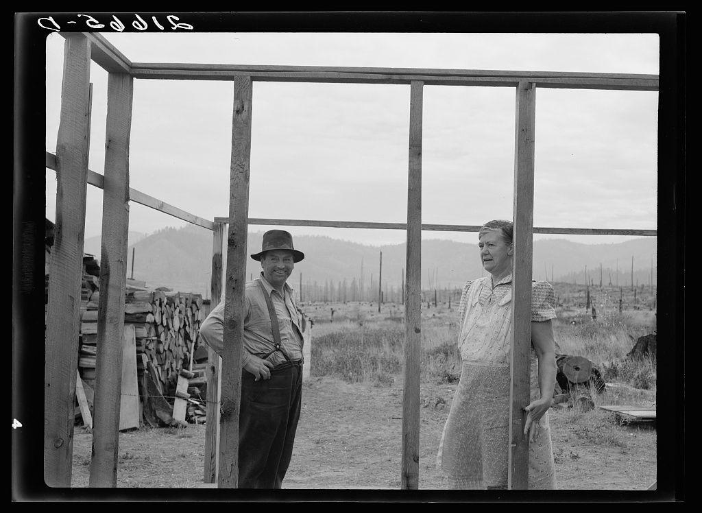 The image captures a moment from the early days of farming, likely in Idaho's Priest River Valley. It shows two individuals engaged in constructing an addition to their one-room cabin on cut-over land. The man is dressed casually with suspenders and a hat while standing confidently next to wooden beams that form part of his new building project. He has his hands resting on the structure, exuding confidence or perhaps fatigue from the manual labor involved.

Standing beside him is another person clad in a dress apron, indicative of domestic tasks often associated with women during this period. The attire suggests she may be responsible for household chores alongside farming duties. Both individuals appear to be focused and engaged in their work environment. In the background, evidence of agricultural activity can be seen through scattered tools and remnants of cleared land, reflecting a bustling yet hardworking rural setting.

The photograph is black-and-white, emphasizing textures rather than colors, which might have been intentionally chosen by Dorothea Lange or was due to technological limitations at that time. The focus on everyday laborers brings attention to the resilience and dedication required in farming life during those times.