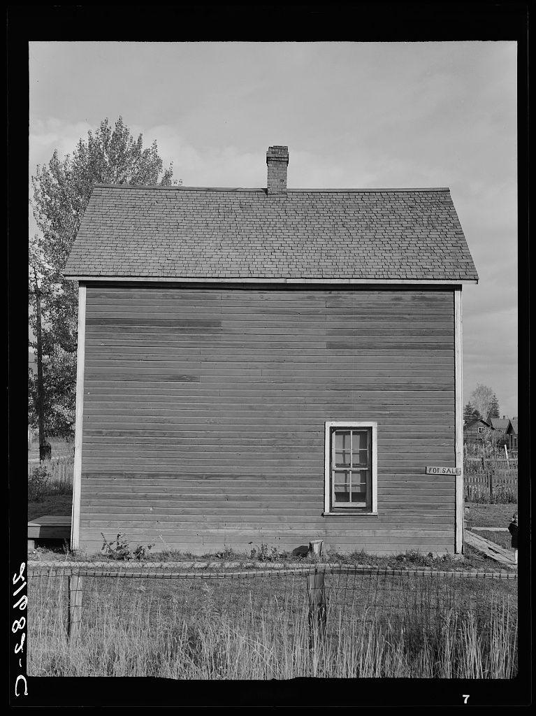The image is a black and white photograph of an old, weathered building with horizontal siding. It appears to be a two-story structure with a steeply pitched roof covered in what seems like shingles or asphalt roofing material. There's no visible snow on the ground, suggesting that it might not currently be winter.

On one side of the house is a small window without any curtains or blinds and another sign attached below this, which appears to read "FOR SALE." The building is situated outdoors with tall grasses surrounding its base, indicating an absence of lawn maintenance. There's also a clear sky above, although it seems slightly overcast.

The ground around the building looks dry but not recently maintained as indicated by weeds and unkempt vegetation growing at some spots near where the house meets the grassy field. In the background to the right, there is another structure which could be part of this property or nearby properties indicating that these buildings are in a rural area with other structures close together.

The overall condition suggests neglect but it's not dilapidated and seems structurally intact despite its old age.