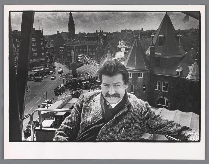 The image is a black and white photograph depicting an individual standing in front of a bustling urban landscape. The person, who appears to be Karel Appel based on the context provided by the reference link, has distinctive features such as wavy hair and a mustache, wearing what seems like a tweed jacket over a dark vest or shirt.

Behind him is a vibrant scene with buildings that have steeply pitched roofs characteristic of European architecture. The skyline includes various structures suggesting an urban environment, possibly in Amsterdam given the reference to "Amsterdam" in the provided link. There's also visible activity on streets and sidewalks below, indicating a lively city atmosphere.

The photograph has been taken from what appears to be another elevated vantage point, perhaps inside or near the ride that Karel Appel is seen beside—a Ferris wheel with multiple gondolas. The angle of capture suggests this might have been taken while riding it or right after exiting. 

There's a juxtaposition of urban life and amusement park elements within the frame, creating an interesting contrast between everyday cityscape and recreational activity.

The photo carries historical significance as indicated by its monochrome quality typical for photographs from earlier decades and serves as visual documentation from around 1961 based on other sources' contextual clues.
