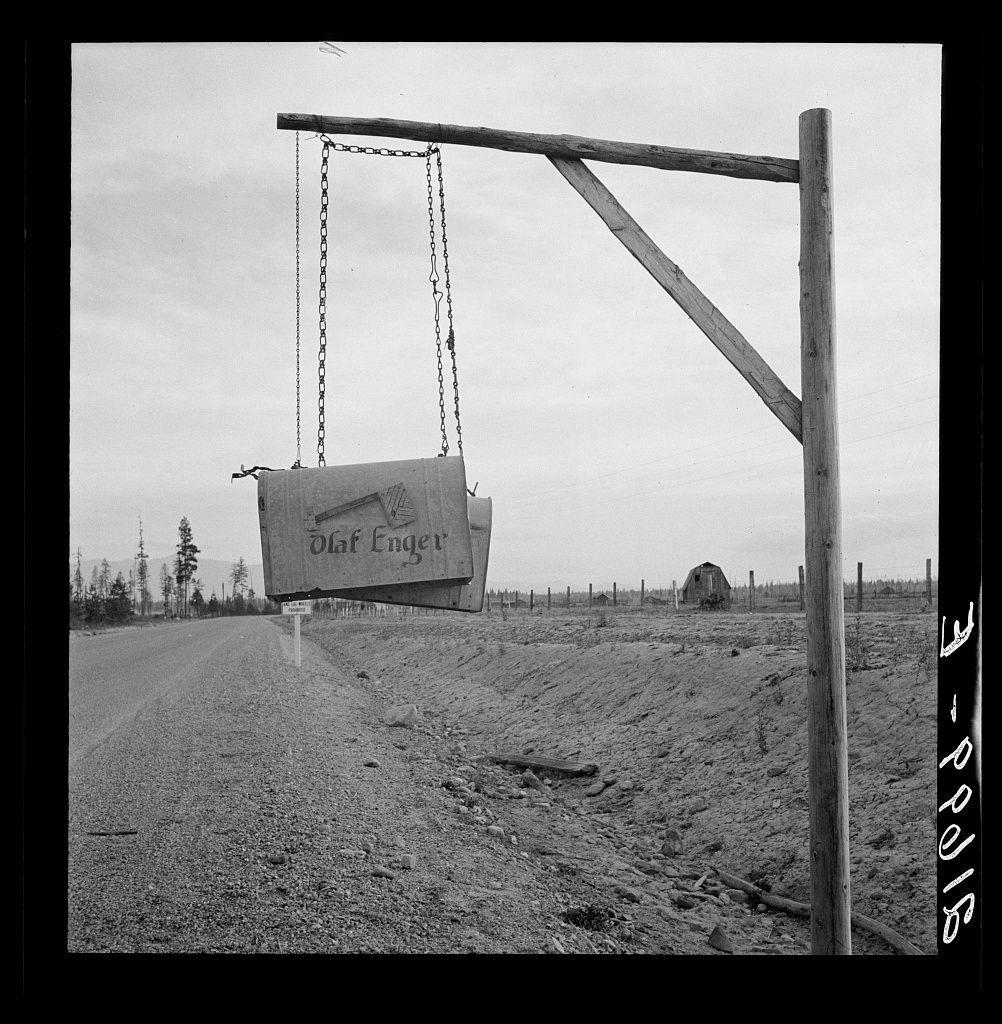 The image captures a monochrome scene depicting an outdoor setting with various elements. In the foreground, there is a piece of heavy machinery suspended in mid-air by chains attached to a metal beam structure. The equipment appears aged and worn out, suggesting it may be part of some construction or maintenance work.

To the right stands a wooden utility pole extending vertically from its base into the upper portion of the frame. Its rustic appearance adds an element of rural or agricultural ambiance to the scene. 

In the background, there is a road that cuts across the landscape, flanked by sparse vegetation and trees in varying distances from each other. The horizon line reveals faint silhouettes of mountains under a sky with scattered clouds.

The overall setting appears somewhat desolate, with no visible human activity or wildlife. It evokes feelings of solitude amidst nature's vast expanse and hints at the challenges faced due to extreme weather conditions like deep snow during winters as mentioned in the source provided (Boundary County, Idaho).
