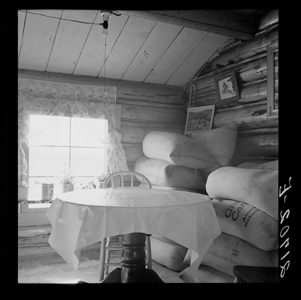 A black and white photograph depicting an interior scene of a rustic, wooden log cabin. The room is furnished with simple furniture including a round table covered in a light-colored cloth draped over it, accompanied by several chairs; one visible on the right side. On the left wall hangs a lace curtain partially obscuring part of a window letting natural light into the space.

Stacks of large burlap sacks are arranged near a corner against the wooden walls, suggesting storage for farming supplies such as grain or seeds. A small potted plant sits next to an old-fashioned lantern on a windowsill. The ceiling is a white-painted plank with some visible cracks and marks indicating age and use.

Various framed photographs adorn one wall of the cabin's interior, adding a personal touch. Above this area hangs what appears to be a simple lamp or light fixture. Overall, the setting suggests an agricultural lifestyle, possibly from mid-20th century America, as indicated by the grain sacks labeled with text such as "Great Northern", which implies commercial farming activity and price fluctuations in commodity markets.

The black-and-white palette emphasizes texture contrasts and historical ambiance without color distraction, providing a timeless snapshot of rural life.