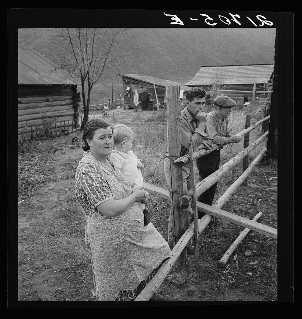 The image is a black and white photograph depicting four individuals, likely from the early to mid-20th century given its vintage appearance. In the foreground, there's an older woman wearing a patterned dress with what appears to be a protective apron over it; she has her arm around a young child who seems slightly uncomfortable or resistant. She is holding another infant in her left hand and looking directly at the camera.

Behind them stands a wooden fence that runs along their property line, separating this area from others beyond which are visible more structures typical of rural farm settings—huts with corrugated metal roofs and exposed beams supporting simple construction. In the midground on the right side is an older man leaning against one section while another man in front of him has his hand under his chin; both men wear casual attire consistent with what might be expected for manual labor.

The setting suggests a rural farm environment, possibly during hard times as indicated by their worn clothes and modest living conditions. The presence of these individuals is supported through the Farm Security Administration (FSA) label on this photo which was an organization under Franklin D. Roosevelt's New Deal programs aimed at helping American farmers in distress due to economic difficulties.

The specific location within Idaho, USA—Priest River Valley—is also indic [...]