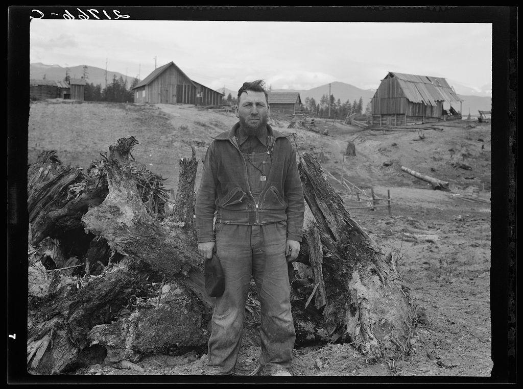 The image is a black and white photograph depicting an individual standing in front of a large, uprooted tree stump. The person appears to be dressed for cold weather or outdoor labor; they're wearing what looks like heavy-duty work clothing: a jacket with visible pockets and buttons, pants that seem sturdy enough for manual tasks, and boots. They are holding something small and dark-colored in their left hand near the waist level.

In the background, there is an open landscape which includes several rustic wooden structures resembling barns or farm buildings, indicative of rural farmland setting. The architecture suggests a historical context with unpainted wood visible on what seems to be weathered walls and roofs. Beyond this immediate scene are mountains in the distance under a cloudy sky.

The ground appears barren and covered with dry grass, which implies an arid environment typical for agricultural regions or post-harvest period where crops have been harvested but not yet replanted. The overcast skies suggest it could be early morning or late afternoon. There is no visible greenery that would indicate active farming in this particular moment captured by the camera.

The watermark on the photo reads 'Dorothea Lange,' which suggests she was a photographer, and her work often documented rural life during times of hardship like the Great Depression for Mennonite farmer [...]