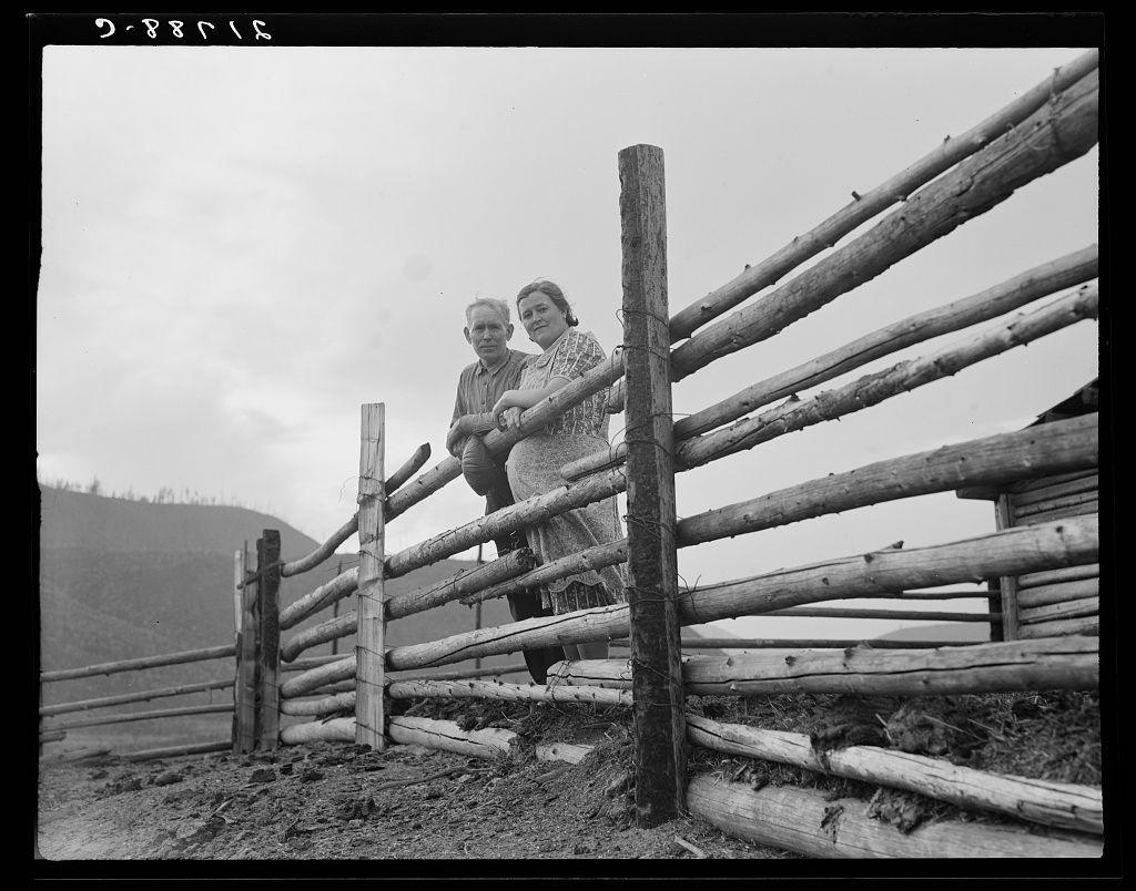 The image is a black and white photograph depicting two individuals, presumably an older couple, standing behind a rustic wooden fence. The man on the left appears to be in his late 60s or early 70s with gray hair, wearing casual work clothes including trousers and a long-sleeve shirt. He has one arm resting on top of the fence while leaning slightly forward.

The woman beside him seems around her mid-50s or older, dressed similarly to the man in practical attire suitable for rural labor; she wears what appears to be a patterned blouse with sleeves rolled up and trousers tucked into boots. Her posture is upright as she stands next to the man, also leaning on the fence.

The background shows an open landscape that suggests a hilly or mountainous region, which could indicate they are in a remote area such as Idaho's Priest River Peninsula mentioned in the caption text. The sky appears overcast, and there isn't much vegetation visible around them except for what looks like sparse grass on the ground near their feet.

The fence is constructed from horizontal logs supported by vertical posts at intervals. It seems to be part of a boundary or enclosure meant to contain livestock or mark property lines typical in rural farming areas. The overall composition, attire, and setting evoke an impression of agricultural life during perhaps early to mid-20th century America, possibly co [...]